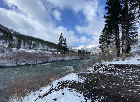 An image of a river flowing thorugh a mountain canyon dusted with snow and pine trees on either side under a partly cloudy blue sky. 