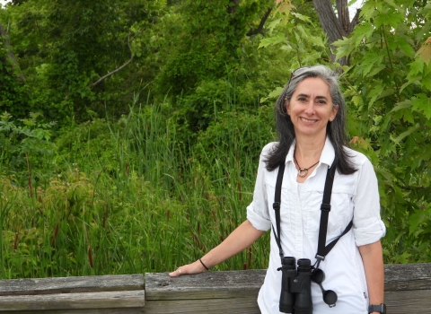 A smiling women with binoculars around her neck wearing a white shirt with greenery in the background