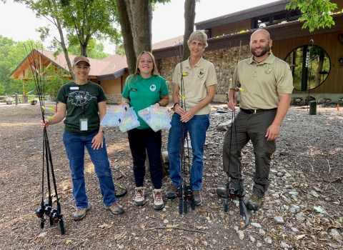 Four adults standing outside next to a building while holding fishing poles and tackle boxes.