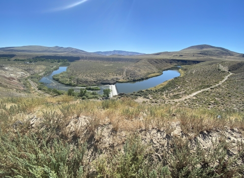 A river running through a desert sage landscape with small mountains in the backdrop and bright blue skies above.