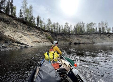 Tim Ericson, sitting at the front of a boat, paddles on the Deshka River. The photo is taken from the back of the boat, and Ericson looks back at the camera over his shoulder. The boat is filled with supplies, including moose antlers. 