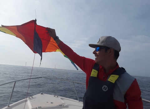 A person wearing a red shirt, black flotation vest and tan cap holds a yellow, red and orange kite aloft while standing in the bow of a boat