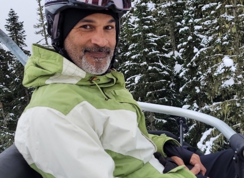 A man smiles while sitting on a ski lift wearing a helmet and green and white jacket. Trees with snow are in the background. 