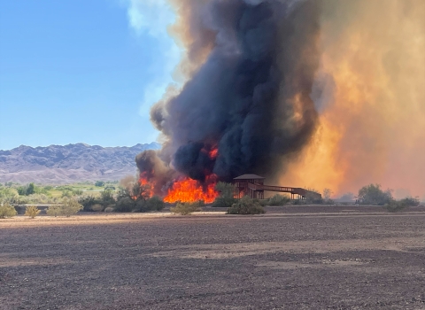 A wildfire is burning in the desert. A black plume of smoke is behind an observation towet