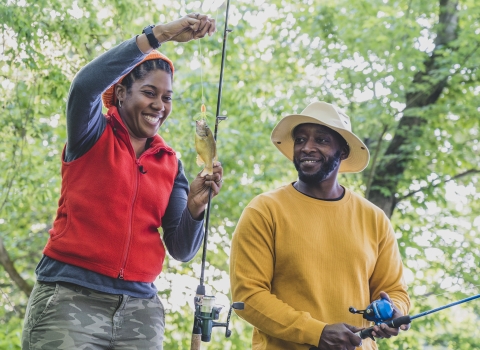 woman reels in small fish while man watches intently 