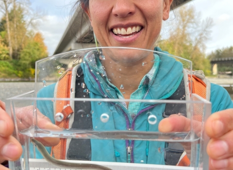 A woman smiles while holding up a clear container holding water and a small, thin eel like fish. The ruler inside measures it around 12 cm in length