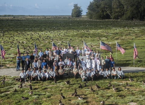 Veterans and Midway Atoll staff pose on field at Midway Atoll surrounding Midway Atoll memorial surrounded by albatross