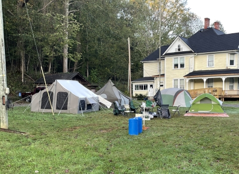 Looking across a green lawn with trees in the background is a large yellow house with a wrap around porch. In the foreground, near the house are several tents set up providing a place for storm volunteers to stay. 