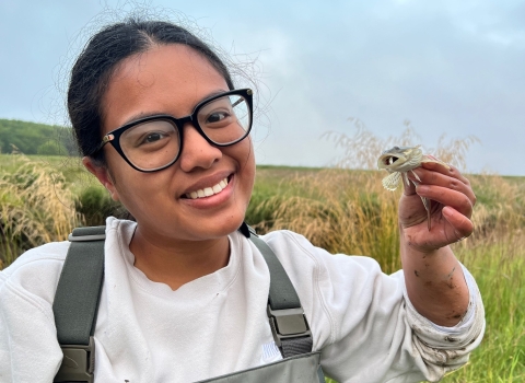 A Pathways employee smiles as she holds up a prickly sculpin. A field and sky can be seen in the background. 