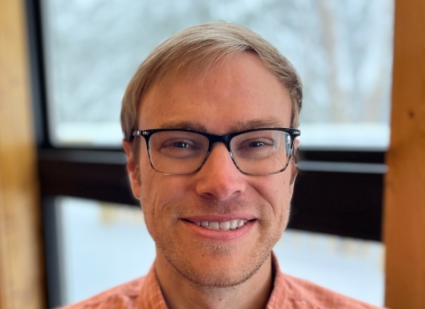 A man with light hair and glasses wearing an orange collared shirt smiles. Behind him is a window.