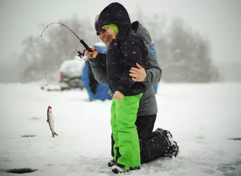 A woman and young boy stand on a frozen lake. The woman is helping the boy hold up a fish they have just caught from a hole in the ice.