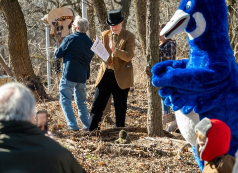 Two men, one holding a guitar, sing next to a blue goose mascot.